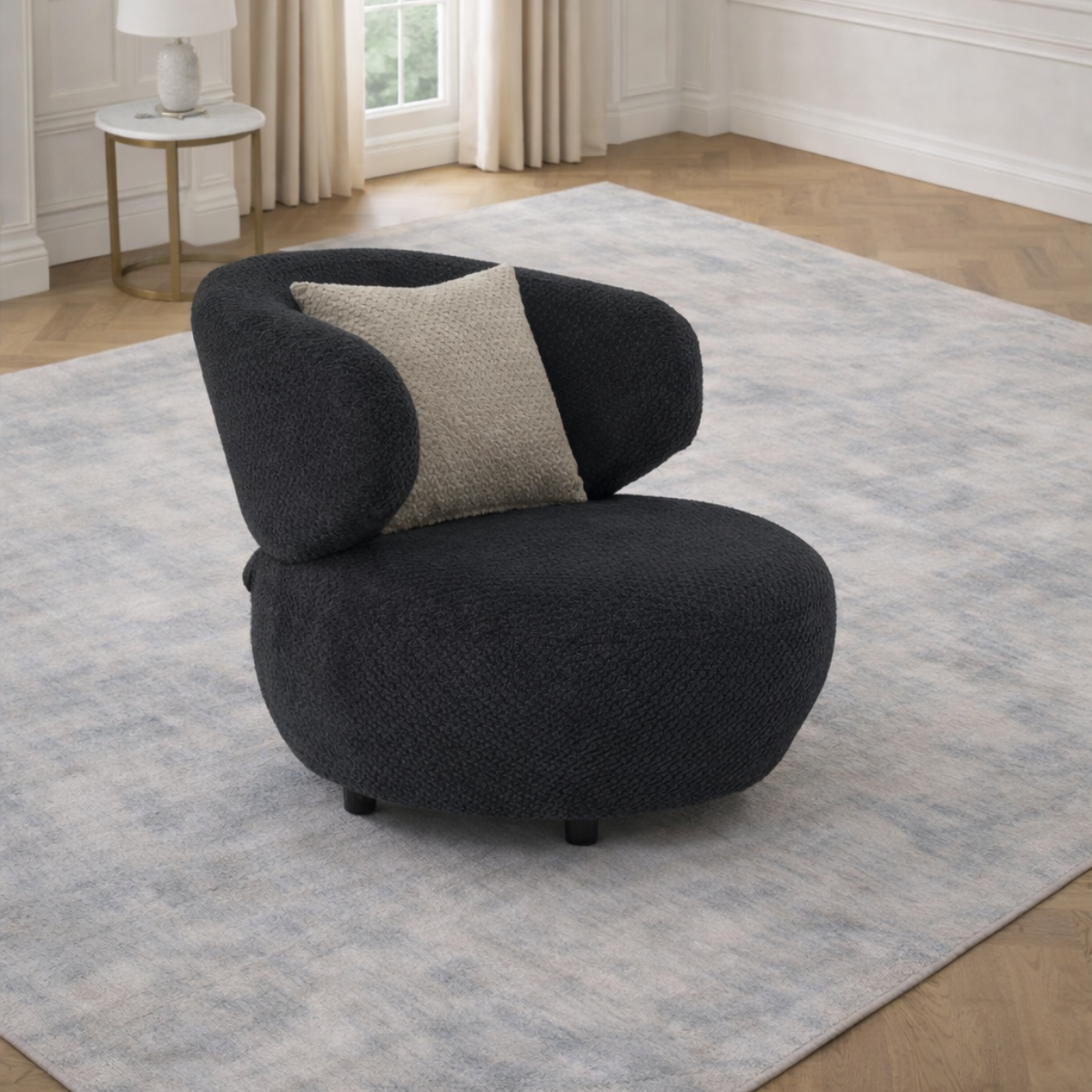 Modern black armchair with a gray pillow on a light blue rug in a room with a window and side table.